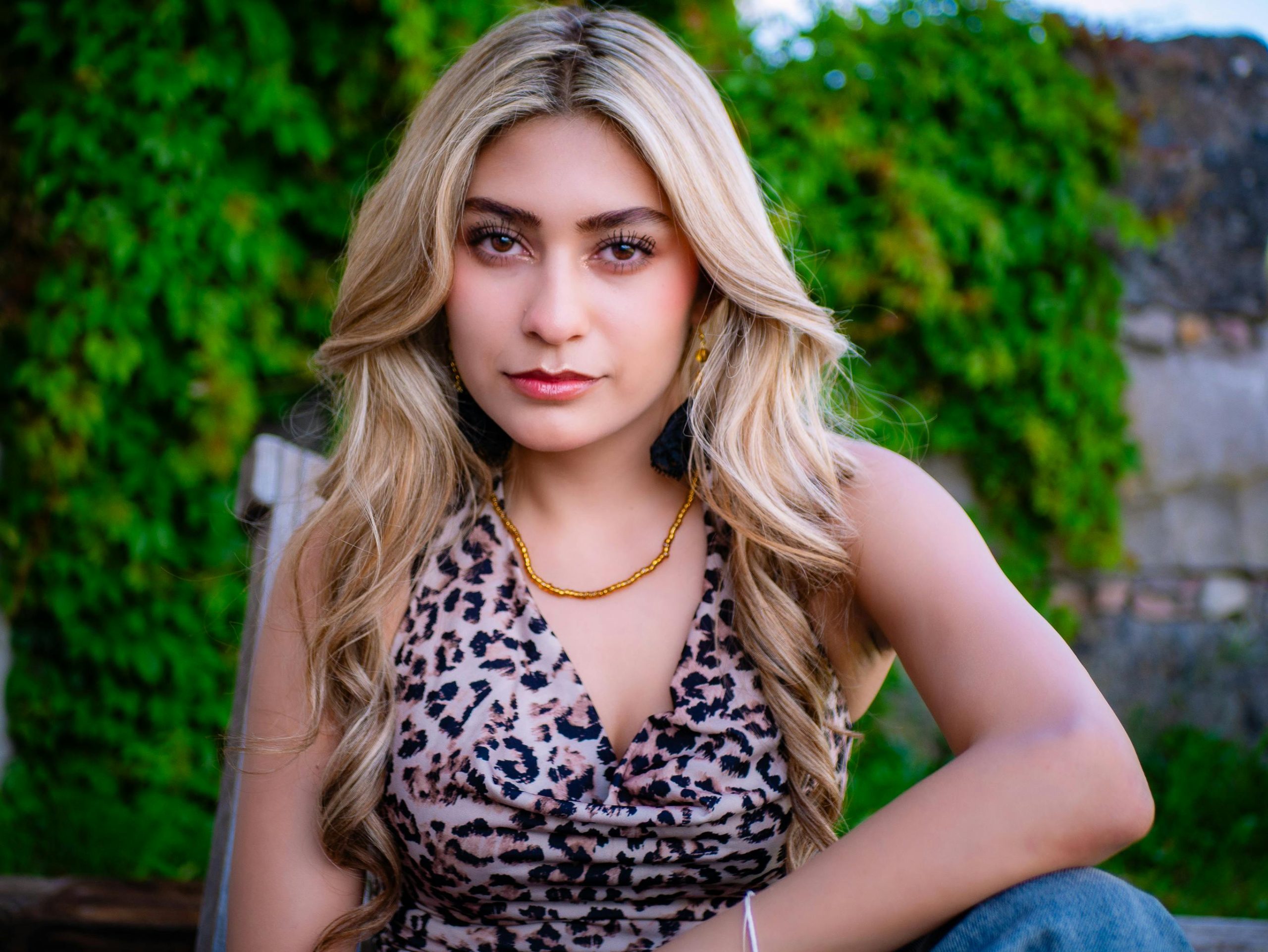 Woman with long blonde hair in a leopard print top against a lush green background, exuding confidence.